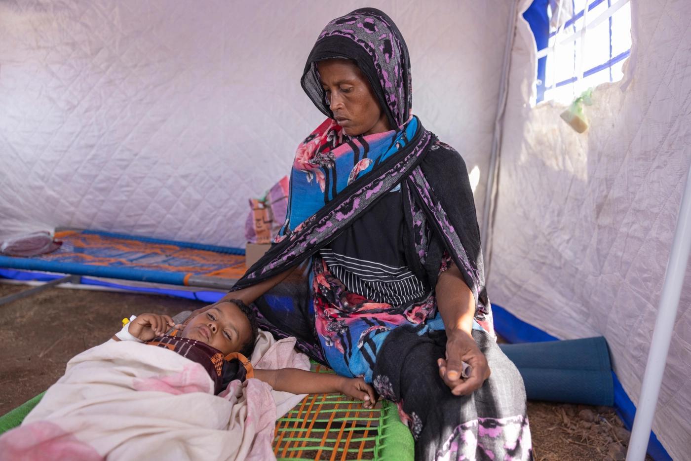 Kvinna sittande på en brits bredvid sjukt barn i flyktingläger