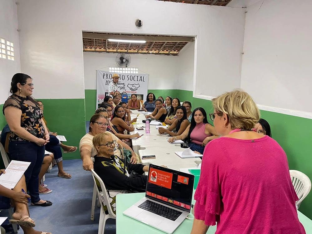 En grupp kvinnor sitter samlade runt ett långt bord i ett färgglatt rum med gröna väggar. De deltar i en workshop eller utbildning ledd av en kvinna i rosa tröja, som står vid ett bord med en bärbar dator. På väggen i bakgrunden syns en affisch med texten "Projeto Social". Stämningen är fokuserad och engagerad.