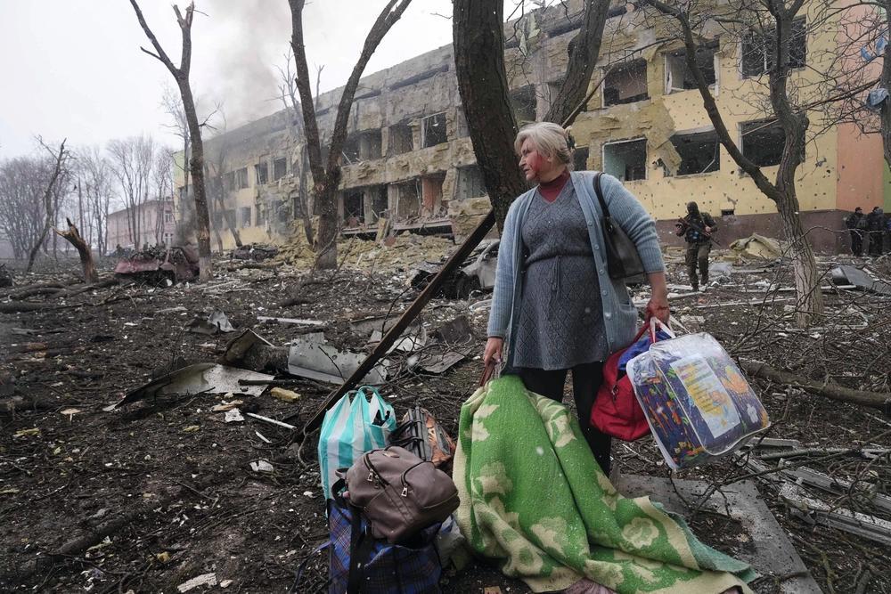 Kvinna med väskor står framför en svårt skadad byggnad och förstörd omgivning efter en attack.